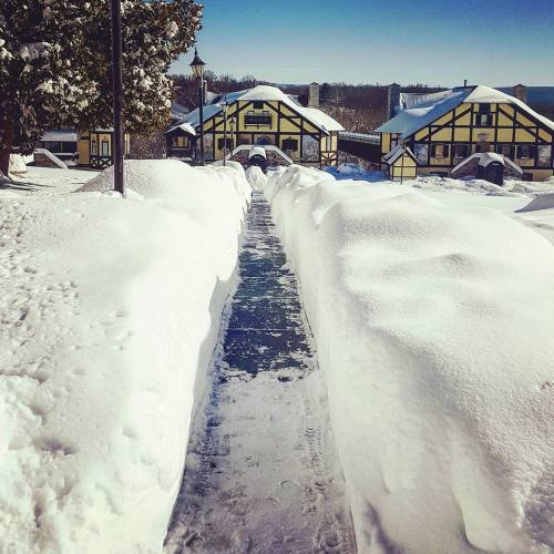 A snowy pathway leads to a yellow and black building, surrounded by deep snow and clear blue skies.