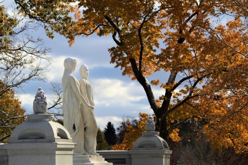 Two white statues stand amidst vibrant autumn trees under a cloudy sky, creating a serene outdoor scene.
