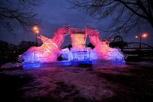 Illuminated ice sculpture of two birds with a sign, set against a twilight sky and surrounded by snow.