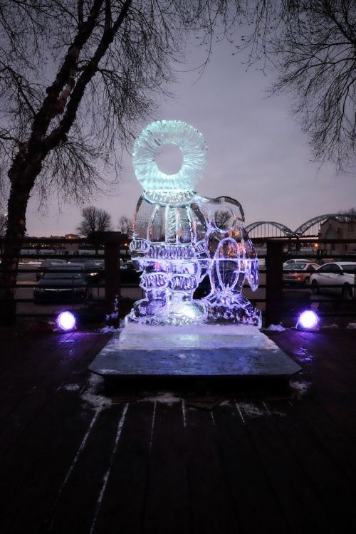 An illuminated ice sculpture of a figure holding a circular object, set against a twilight sky and surrounded by trees.