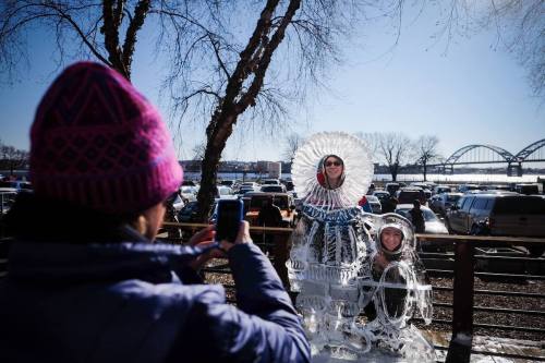 A person takes a photo of another person posing with an ice sculpture on a sunny day, surrounded by trees and cars.