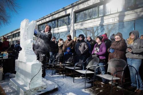A sculptor carves an ice statue while a crowd watches in a snowy outdoor setting.