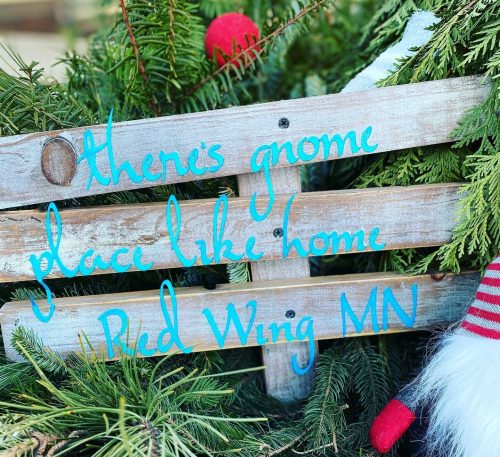 A wooden sign among greenery reads, "There's gnome place like home, Red Wing MN," with festive decorations.
