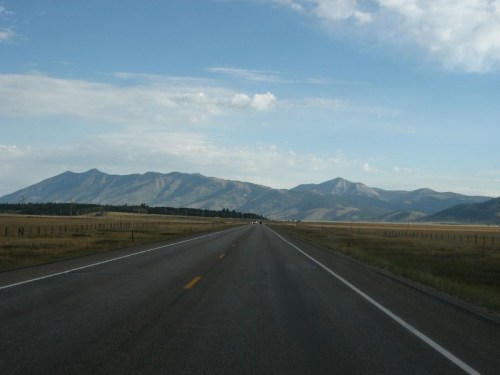 A straight road stretches into the distance, flanked by open fields and mountains under a partly cloudy sky.