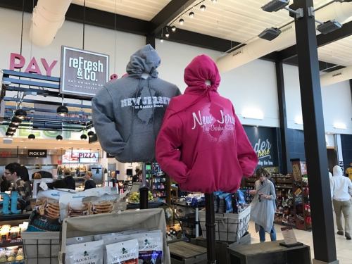 Two hoodies on display, one gray and one pink, featuring "New Jersey" text, in a busy retail space.
