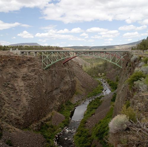 A green and red bridge spans a deep canyon, with a river flowing below and clouds in a blue sky above.
