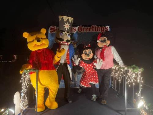 Four costumed characters, including Winnie the Pooh and Minnie Mouse, pose together in a festive setting with holiday decorations.