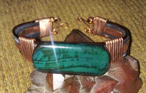 A copper and leather bracelet featuring a large green stone, resting on a textured surface.