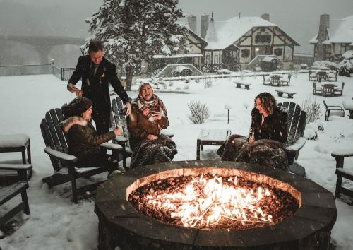 A group of four people enjoying drinks by a fire pit in a snowy outdoor setting, with a cozy lodge in the background.