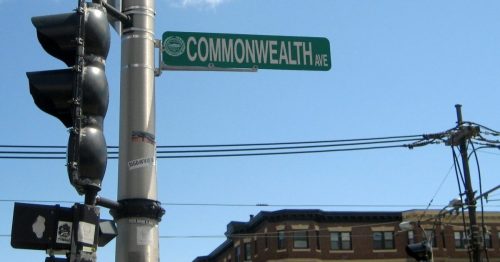 Street sign for Commonwealth Avenue against a clear blue sky, with traffic lights and buildings in the background.