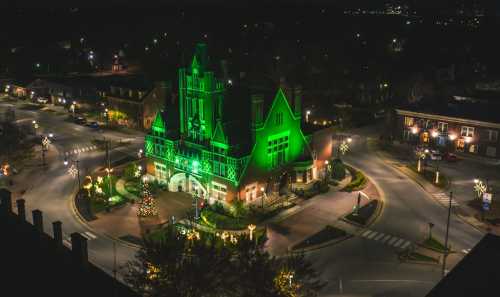 Aerial view of a green-lit building surrounded by festive lights and a Christmas tree in a charming town square at night.