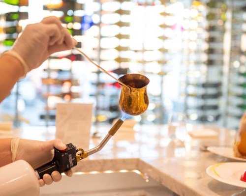 A person in gloves uses a torch to heat a small golden pot in a brightly lit kitchen with colorful glass in the background.