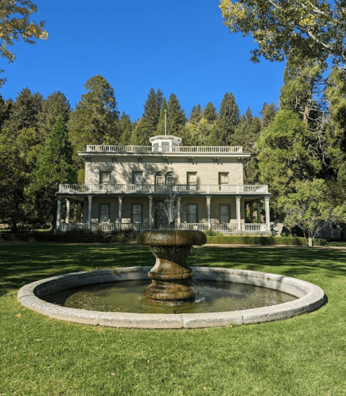 Historic mansion with a fountain in the foreground, surrounded by lush greenery and tall trees under a clear blue sky.