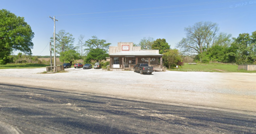 A small country store with a gravel parking lot, surrounded by trees and open fields under a clear blue sky.