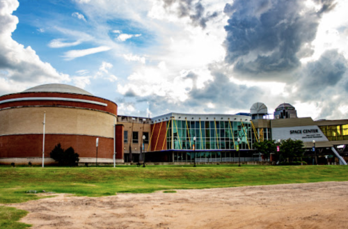 A modern space center building with a dome, large glass windows, and a grassy area under a cloudy sky.