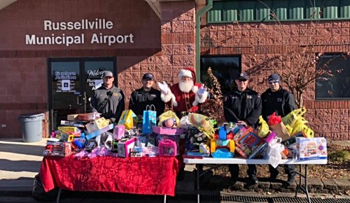 A group of five people, including Santa, stands by a table filled with toys outside Russellville Municipal Airport.