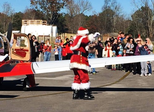 Santa Claus waves to a crowd gathered near a small airplane on a sunny day, with festive decorations in the background.