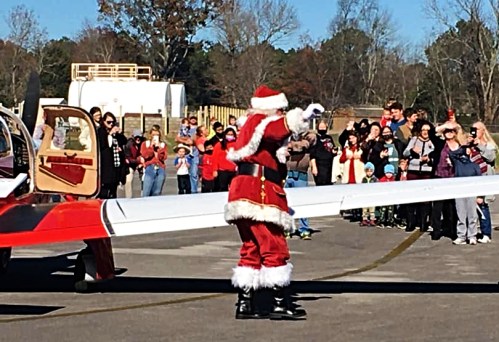 Santa Claus waves to a crowd of people gathered near a small airplane on a sunny day.