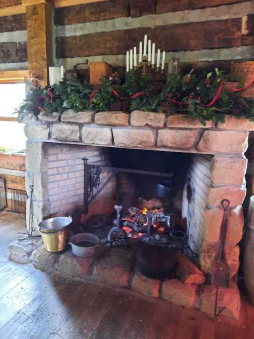 A rustic stone fireplace adorned with greenery and candles, featuring a warm fire and various cooking pots.