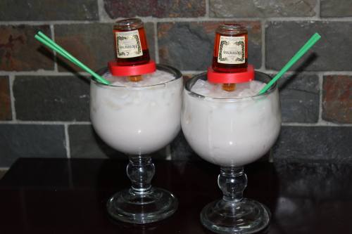 Two large glasses filled with a creamy drink, each topped with a small bottle and green straws, set against a stone background.