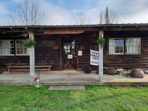 A rustic wooden building with a sign reading "Frog Trading Post Open" and potted plants on the porch.