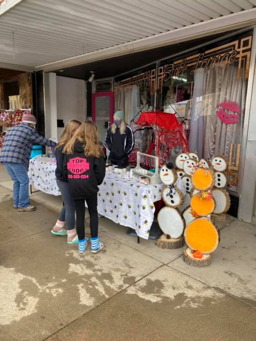 Two girls browse a vendor's table with crafts, while a woman assists them outside a shop decorated with snowmen.