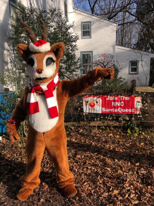 A person in a reindeer costume with a scarf points towards a festive sign in a winter setting.