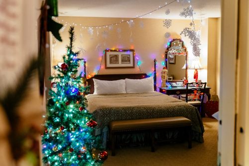 Cozy hotel room decorated for Christmas, featuring a bed, colorful lights, a small tree, and festive ornaments.