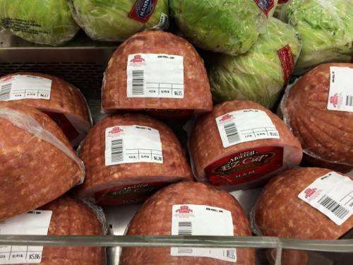 Several packaged hams with price tags, placed among fresh heads of lettuce in a grocery store display.