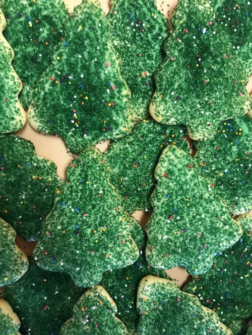 A close-up of green Christmas tree-shaped cookies covered in sparkling sugar and colorful sprinkles.