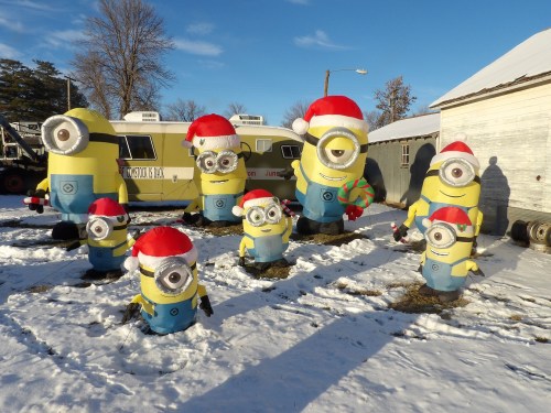 A group of Minion figures wearing Santa hats in a snowy outdoor setting, with a yellow trailer in the background.