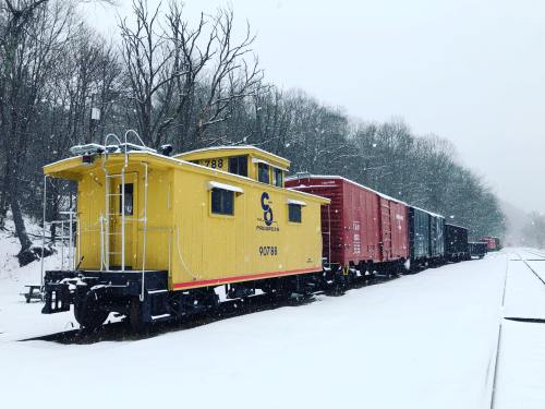 A yellow caboose and several freight cars sit on a snowy train track surrounded by trees. Snow is falling gently.