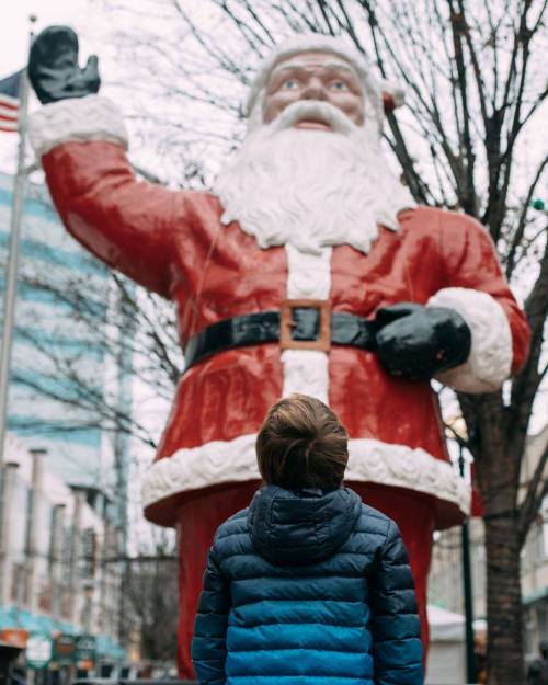 A child in a blue jacket stands in front of a large Santa Claus statue, waving in a festive outdoor setting.