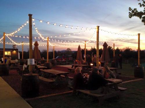 Outdoor seating area with string lights at sunset, featuring picnic tables and umbrellas, with people enjoying the ambiance.