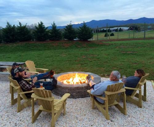 A group of four people relax around a fire pit, seated in wooden chairs, with mountains in the background.