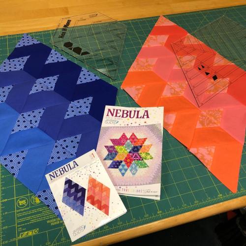 Colorful quilt pieces in blue and pink, with pattern templates labeled "NEBULA" on a green cutting mat.