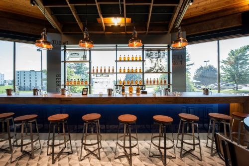 A modern bar with wooden stools, a blue counter, and shelves displaying various bottles against a bright window backdrop.