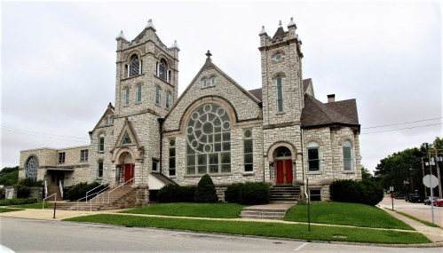 Historic stone church with large stained glass windows and twin towers, set in a grassy area with a cloudy sky.