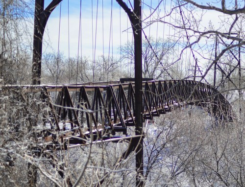 A rusted suspension bridge surrounded by bare trees and snow, set against a clear blue sky.