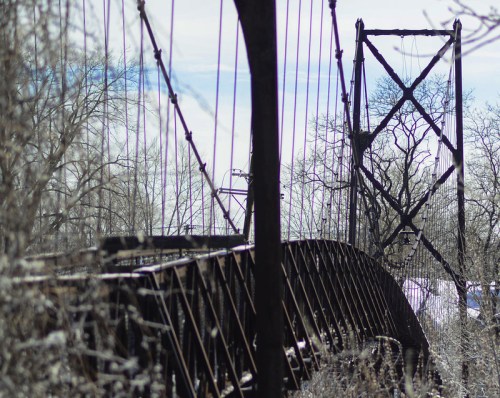 A weathered suspension bridge surrounded by bare trees and a clear sky, showcasing intricate metalwork and cables.