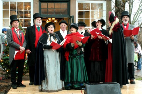 A group of carolers in Victorian attire singing outdoors, surrounded by festive decorations and a cheerful crowd.