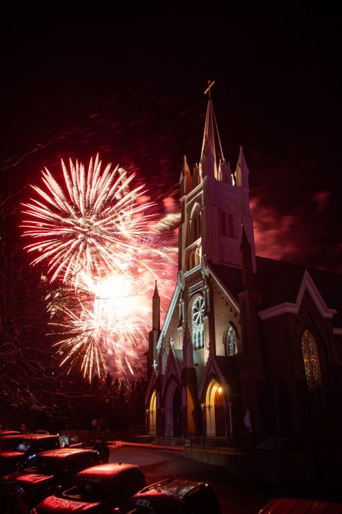 A church illuminated by colorful fireworks in the night sky, with cars parked in the foreground.
