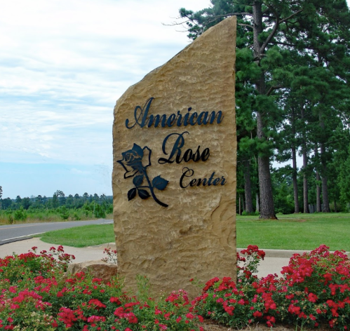Sign for the American Rose Center, featuring a rose design, surrounded by blooming red flowers and tall trees.