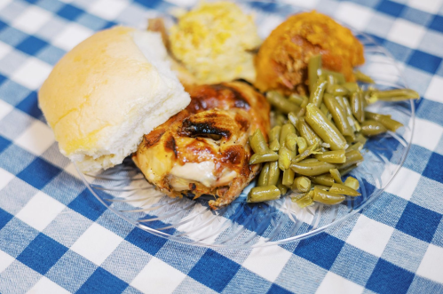 A plate of grilled chicken, green beans, cornbread, and a roll on a checkered tablecloth.