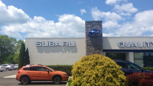 Signage for a Subaru dealership with an orange car parked outside under a blue sky with fluffy clouds.