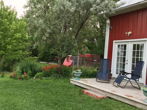 A red barn-style house with a porch, two chairs, and lush greenery, including flowers and trees in the background.
