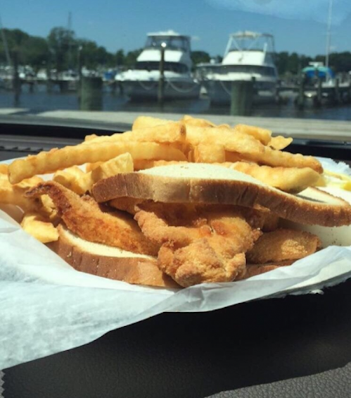 A plate of fried fish and crinkle-cut fries served on bread, with boats in the background by a marina.