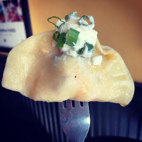 A close-up of a pierogi topped with sour cream and green onions, held on a fork against a warm background.