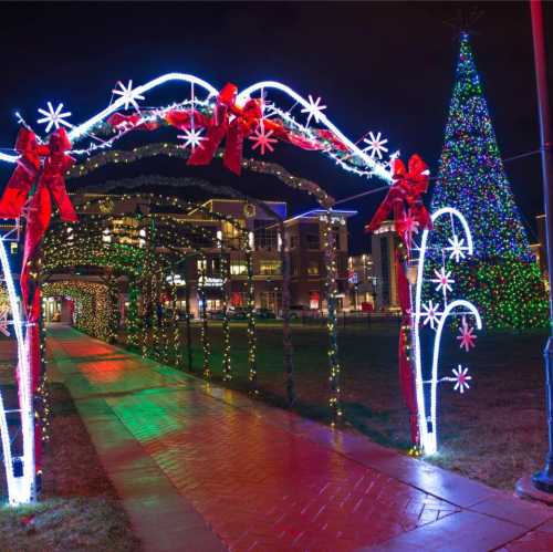 A festive pathway adorned with colorful lights and bows, leading to a large, illuminated Christmas tree.