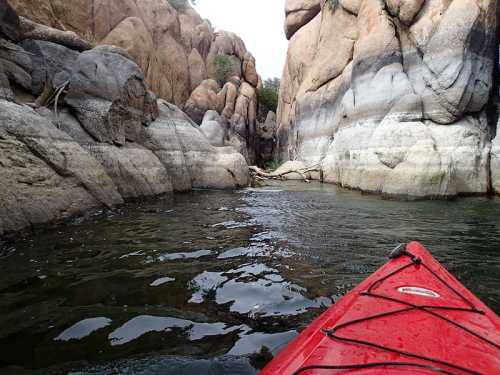 A red kayak glides through a narrow canyon with towering rock formations and calm water.